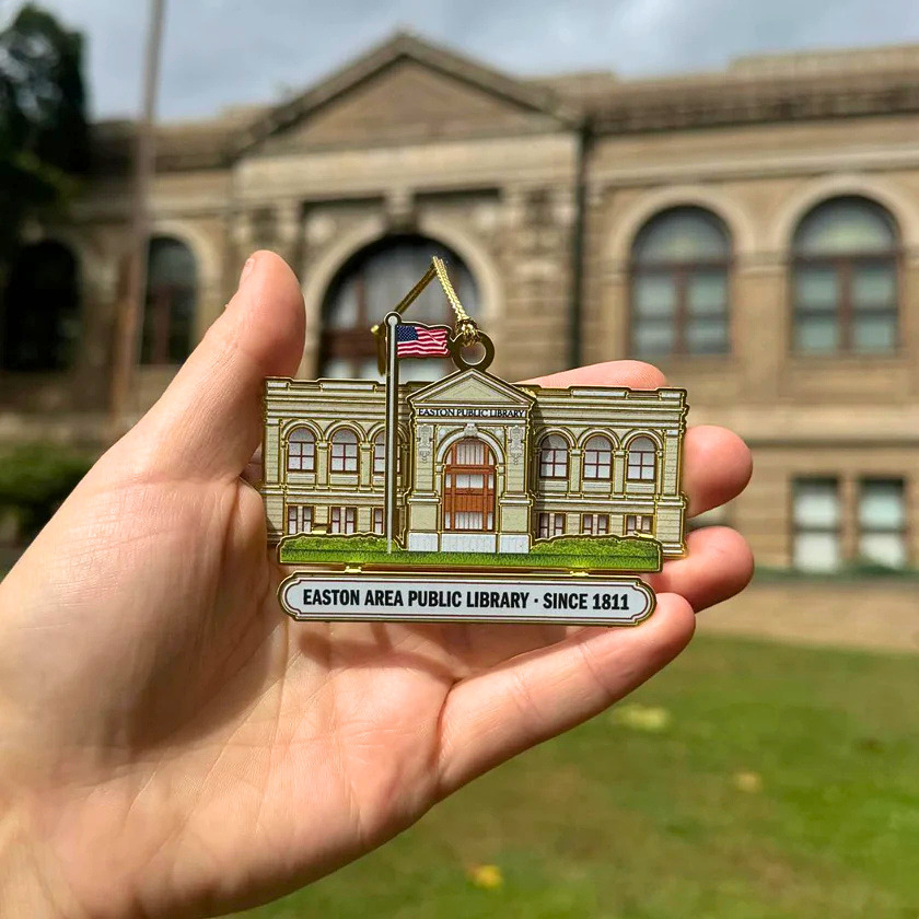 A hand holding a detailed ornament of the Easton Area Public Library in front of the real library building, with the facade softly blurred in the background.