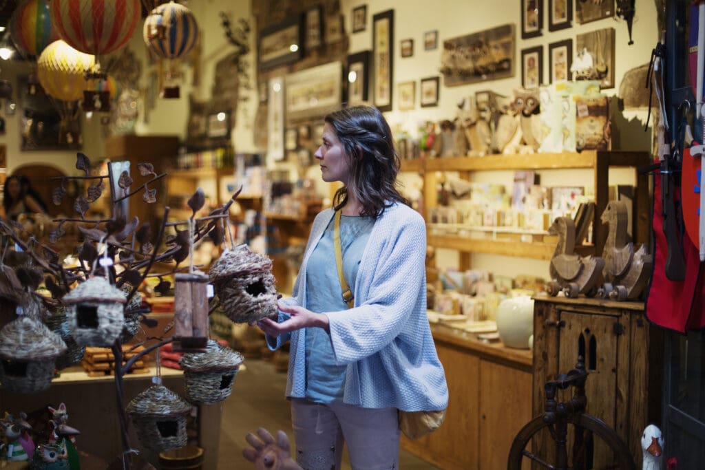 Customer browsing decorative items inside a gift shop, holding a small woven birdhouse ornament near a display stand.